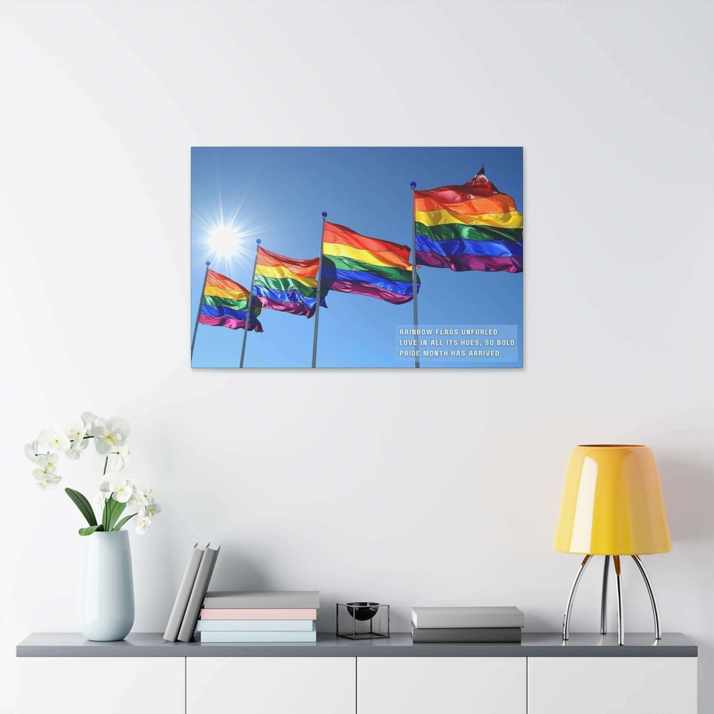 A collection of Bold Rainbow Flags Canvas Wall Art from Printify is shown flying in the breeze against a clear blue sky, with sunlight shining brightly in the background. The text on the vibrant Pride Month Art Print reads, "RAINBOW FLAGS UNFURLED LOVE IN ALL ITS HUES, SO BOLD PRIDE MONTH HAS ARRIVED," celebrating the LGBTQ+ community.